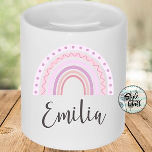 May include: White ceramic money box with a pink, purple and grey rainbow design and the name "Emilia" written in black below the rainbow.