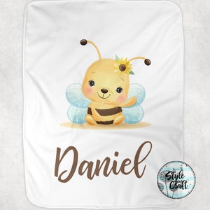 May include: A white baby blanket with a cute cartoon bee wearing a sunflower. The bee is yellow and black with blue wings. The blanket has the name "Daniel" printed in brown text.
