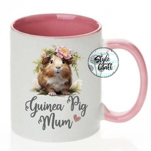 May include: White ceramic mug with a pink interior and handle. The mug features a graphic of a guinea pig wearing a flower crown and the text "Guinea Pig Mum" with a pink heart.