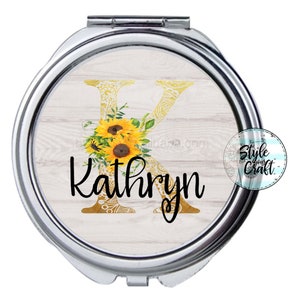 May include: A silver compact mirror with a white woodgrain background. The letter K is gold with a floral pattern and a sunflower wreath. The name Kathryn is written in black below the letter.