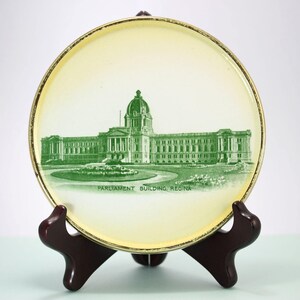 May include: A decorative plate with a green illustration of the Parliament Building in Regina, Canada. The plate has a cream background with a gold rim and is on a wooden stand.