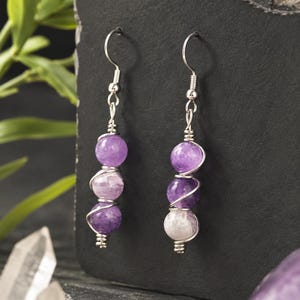 May include: A pair of drop earrings featuring three round purple amethyst beads each, wrapped in silver-toned wire. The earrings hang from silver-toned fishhook ear wires, showcasing natural stone variations in shades of light and deep violet.