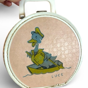 May include: Vintage round lunchbox with a white handle and trim. The pink lid features a cartoon duck in a boat, wearing a sailor's hat and vest. The lunchbox has the text "L-U-C-E" on the bottom right.