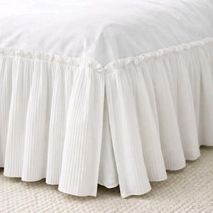 May include: A white bed skirt featuring a pleated design and a ruffled edge. The bed skirt is made of a soft, flowing fabric, adding elegance to the bed. A classic and timeless piece for any bedroom.