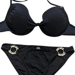May include: Black bikini set featuring a halter top and low-rise bottoms. The top has a knotted design in the center. The bottoms have gold-colored circular accents on each side. The swimwear is made of a smooth, solid black fabric.