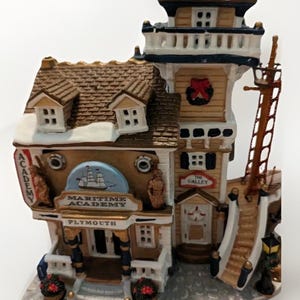 May include: A detailed ceramic Maritime Academy building figurine. The building features a brown roof with snow, a tower, and a sign that reads "The Galley." The front of the building has the words "Maritime Academy Plymouth" and a ship illustration.