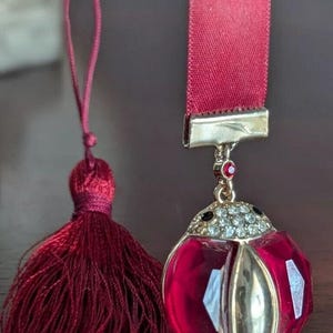 Op de afbeelding: Een rode satijnen bladwijzer met een gouden lieveheersbeestje-bedel. Het lieveheersbeestje heeft een rood lichaam met heldere strass steentjes en zwarte accenten. De bladwijzer heeft aan het ene uiteinde een kwast.