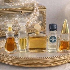 May include: An assortment of antique perfume bottles, each with a unique design and filled with coloured liquids. The bottles are displayed on a gold-coloured tray, with a jewellery box and pearls visible in the background.
