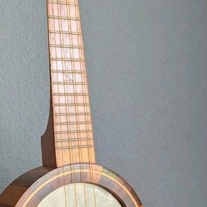 May include: A vintage banjo with a wooden neck and body. The round body features a light-colored membrane and is framed by decorative wood inlays. The instrument has four strings and tuning pegs.