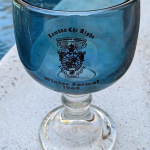 Könnte beinhalten: Ein blaues Glasgefäß mit einer schwarzen Inschrift, die "Lambda Chi Alpha" und "Winter Formal 1969" lautet. Das Gefäß hat einen klaren Glasstiel und eine klare Glasbasis.