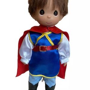 May include: A porcelain doll dressed as a prince, wearing a blue tunic with gold trim, a red cape, and black boots. The doll has brown hair and is standing on a white base with the words "Precious Moments" printed on it.