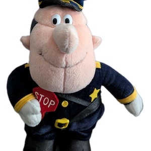 May include: Plush toy of a cartoon traffic officer. The officer wears a navy blue uniform with gold accents, a dark blue hat, and holds a red "STOP" sign. The toy has a smiling face and is made of soft fabric.