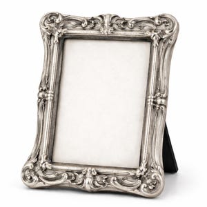 May include: Silver-coloured, ornate picture frame with a blank white space for a photo. The frame has a detailed, scrolling design and a black easel back for tabletop display. The frame is made of a metallic material.