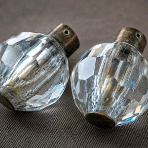 Op de afbeelding: Twee bolvormige kastknoppen met heldere, veelzijdige glazen oppervlakken die licht reflecteren. Elke knop is bevestigd aan een cilindrische, verweerde bronskleurige metalen basis met een klein schroefgat aan de zijkant voor montage.