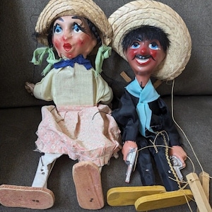 Könnte beinhalten: Ein Paar Vintage-Handpuppen mit Holzbeinen. Die weibliche Puppe trägt einen Strohhut, eine gelbe Bluse, einen rosa Rock und weiße Schuhe. Die männliche Puppe trägt einen Strohhut, eine schwarze Jacke, ein blaues Hemd, eine schwarze Hose und braune Schuhe. Beide Puppen haben bemalte Gesichter und halten Requisiten.