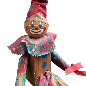 May include: A wooden clown doll with a painted face and a red, blue, and yellow fabric hat and clothing. The doll has a wooden body and blue painted eyes.