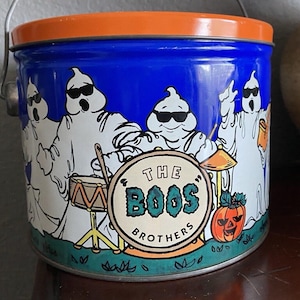 Op de afbeelding: Een blauwe en oranje Halloween-blikken doos met een cartoon-spookband met zonnebrillen. De doos leest "The Boos Brothers" en bevat een pompoen. Het heeft een zilveren handvat.
