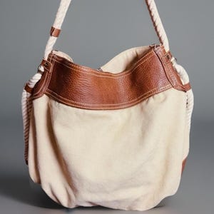 May include: Cream-colored canvas shoulder bag with brown leather trim and accents. The bag features a rope shoulder strap and a spacious interior. The leather trim is stitched with white thread, adding a contrasting detail to the design.
