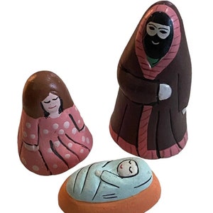 May include: A nativity scene set with three ceramic figurines: a woman in a pink and white polka dot dress, a man in a brown robe with a pink hood, and a baby in a blue blanket lying in an orange crib.