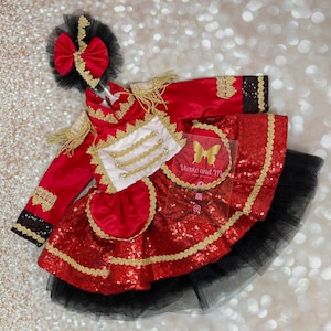 Puede incluir: Un vestido rojo y dorado con lentejuelas, con una falda de tul negro y una chaqueta roja y dorada con ribetes dorados. El vestido tiene una faja blanca con botones dorados y un lazo rojo en la cabeza.