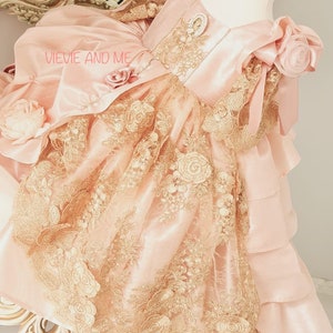 Pode incluir: Um vestido rosa claro com intrincada sobreposição de renda dourada e detalhes florais. O vestido apresenta um corpete de cetim, saia em camadas e enfeites decorativos. O texto "VIEVIE AND ME" é visível.