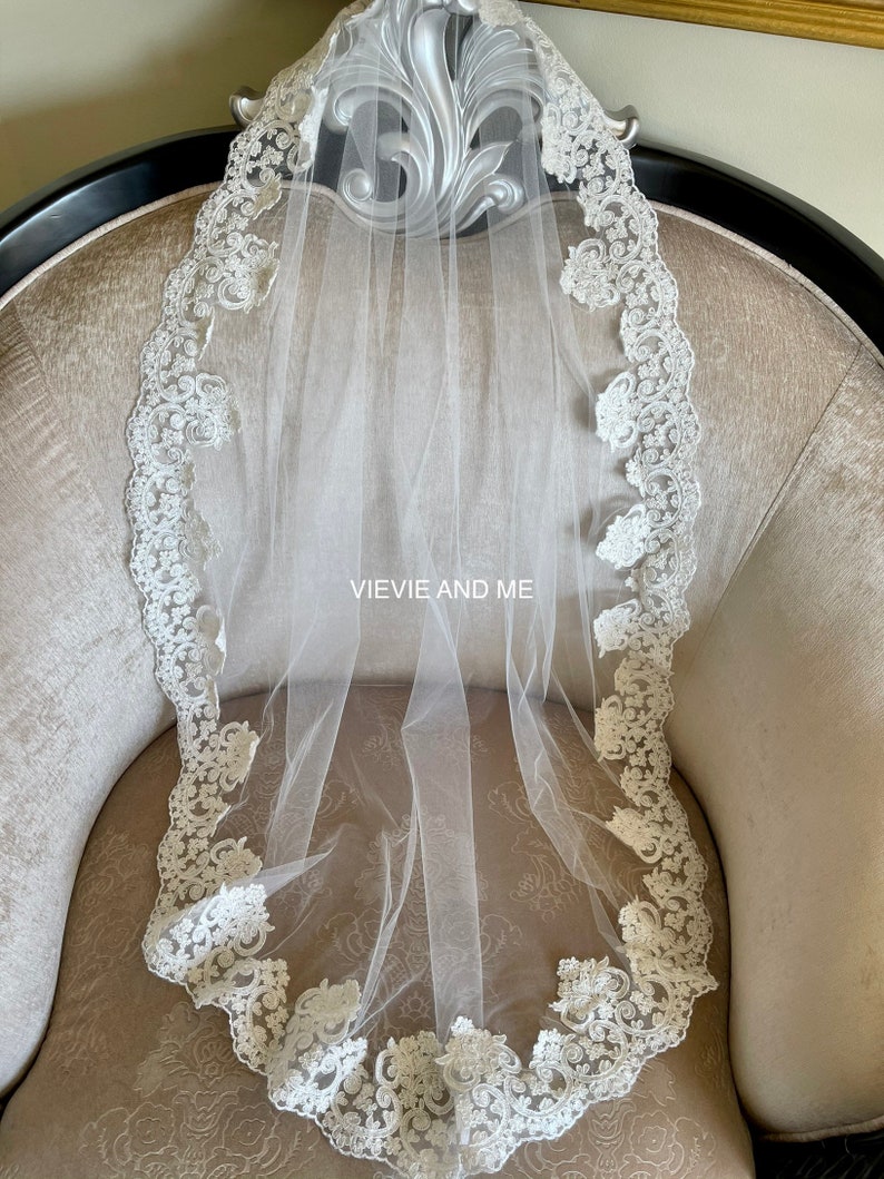 May include: A white bridal veil with a delicate lace trim. The lace features a floral design with a scalloped edge.
