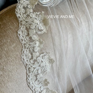 May include: A white bridal veil with a delicate lace trim. The lace features a floral pattern with a scalloped edge. The veil is draped over a chair with the text "VIEVIE AND ME" visible.