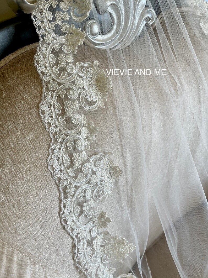 May include: A white bridal veil with a delicate lace trim. The lace features a floral pattern with a scalloped edge. The veil is draped over a chair with the text "VIEVIE AND ME" visible.