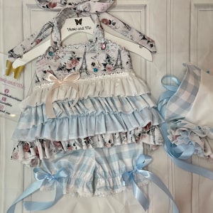 Pode incluir: Um conjunto para menina bebé com estampa xadrez azul e branco. O conjunto inclui uma blusa com babados com estampa floral e da Torre Eiffel, uma calça combinando e um chapéu. A blusa tem uma borda de renda branca e um laço rosa. O conjunto é decorado com pedrarias e um pingente de borboleta. O texto "Weeve and Me" está impresso na blusa.