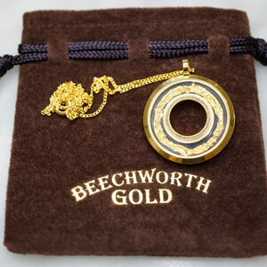 May include: A gold-colored pendant necklace with a circular design, featuring a black center and gold leaf accents. The necklace rests on a brown velvet pouch with the text "BEECHWORTH GOLD" in gold lettering.
