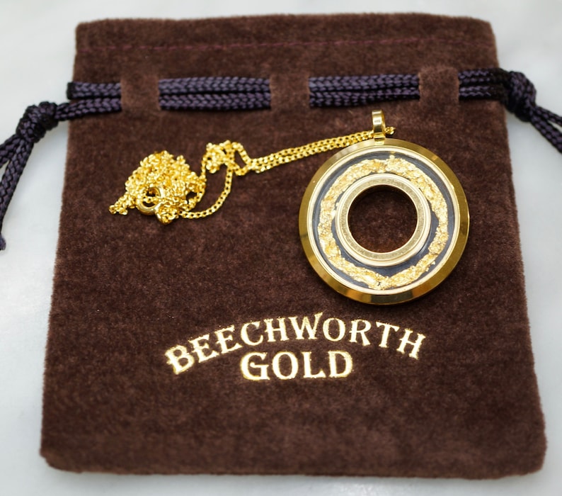 May include: A gold-colored pendant necklace with a circular design, featuring a black center and gold leaf accents. The necklace rests on a brown velvet pouch with the text "BEECHWORTH GOLD" in gold lettering.
