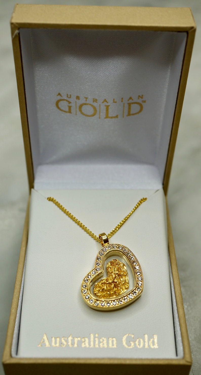 May include: A gold heart-shaped pendant necklace with a clear glass insert containing a piece of Australian gold. The necklace is in a white box with the words "Australian Gold" printed on the lid.
