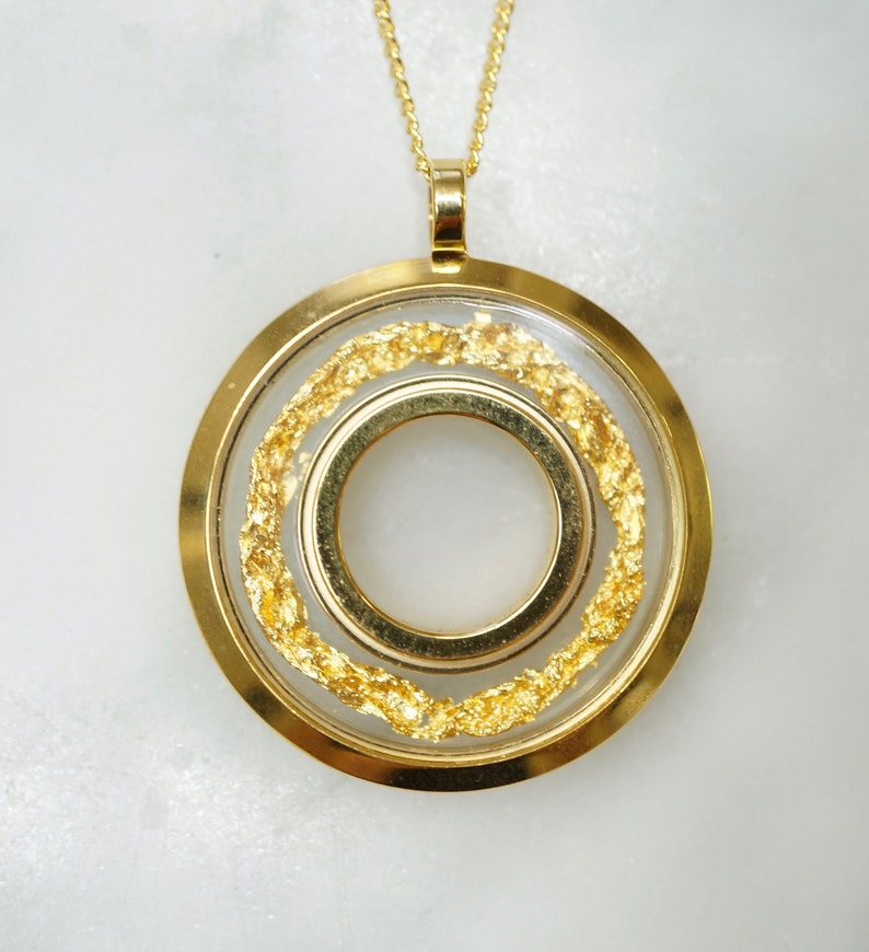 May include: A gold-toned pendant necklace featuring a circular design with a clear center. The pendant has two concentric rings, with gold flakes suspended between the outer ring and a clear inner layer. The necklace chain is also gold.