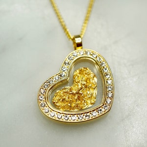 May include: A gold heart-shaped pendant necklace with a clear glass insert containing a piece of gold flake. The pendant is surrounded by a row of clear cubic zirconia stones.