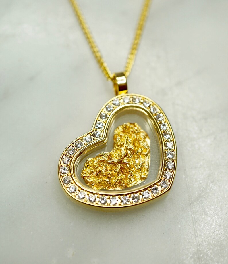 May include: A gold heart-shaped pendant necklace with a clear glass insert containing a piece of gold flake. The pendant is surrounded by a row of clear cubic zirconia stones.