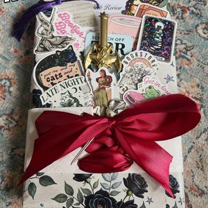 May include: A rectangular package wrapped in floral paper with a large dark red satin ribbon. The package is decorated with various stickers, a gold-colored sword charm, and a purple tassel. The text Romantasy is visible at the top.