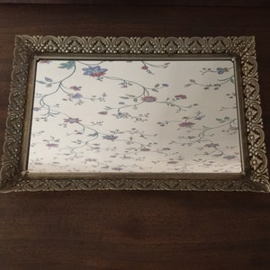 May include: A silver-toned tray with an ornate frame and a mirrored surface. The mirror reflects a floral wallpaper pattern with pink, blue, and green flowers.