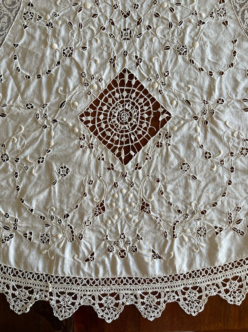 May include: White cotton tablecloth with intricate embroidered lace detailing. The centre features a large, openwork diamond design with a spiral motif. The border is a delicate crochet lace.