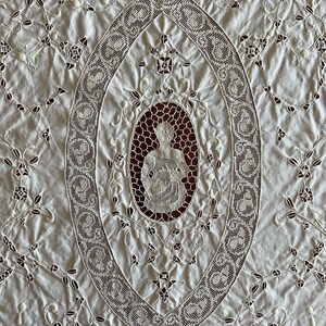 May include: White cotton tablecloth with intricate lace embroidery. The centre features a large oval design with a smaller oval inside depicting a figure holding a bouquet of flowers.