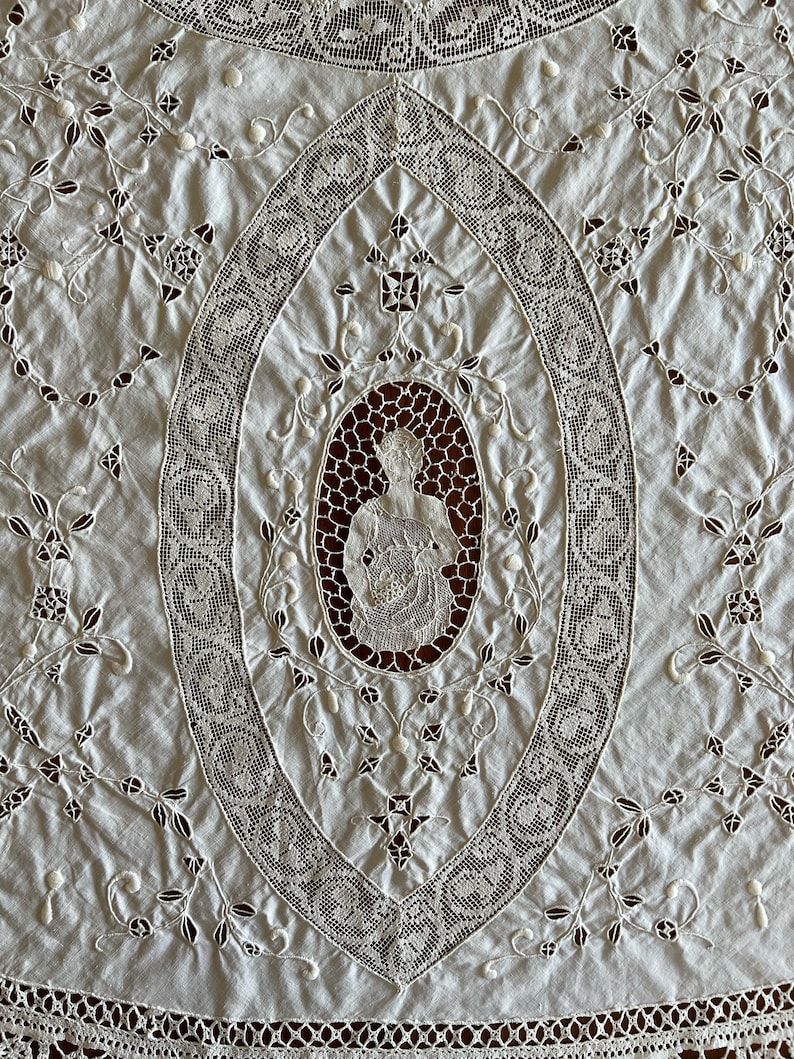 May include: White cotton tablecloth with intricate lace embroidery. The centre features a large oval design with a smaller oval inside depicting a figure holding a bouquet of flowers.
