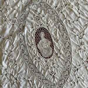 May include: A white cotton tablecloth with intricate lace embroidery featuring a floral design and a central oval with a woman's figure. The tablecloth has a scalloped edge.