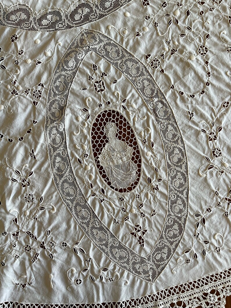 May include: A white cotton tablecloth with intricate lace embroidery featuring a floral design and a central oval with a woman's figure. The tablecloth has a scalloped edge.