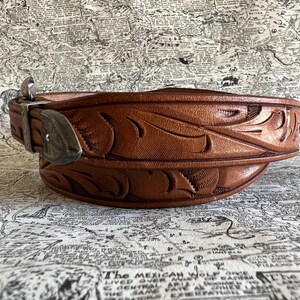May include: A brown leather belt with intricate floral carvings. The belt features a silver-colored buckle and keeper. The leather has a rich, warm tone, and the design is embossed across the entire length of the belt. The belt is a classic Western style.