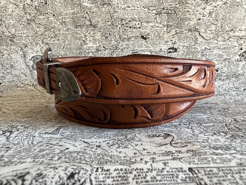 May include: A brown leather belt with intricate floral carvings. The belt features a silver-colored buckle and keeper. The leather has a rich, warm tone, and the design is embossed across the entire length of the belt. The belt is a classic Western style.