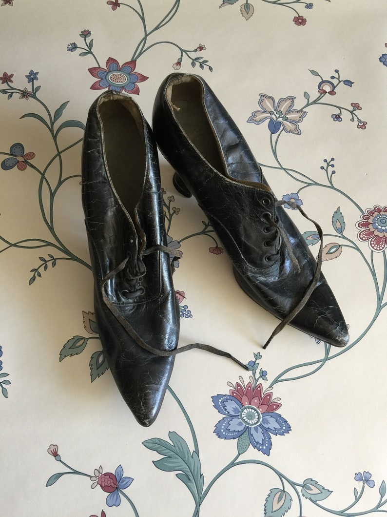 May include: A pair of black leather lace-up shoes with pointed toes and a high heel. The shoes are worn and have a vintage look.
