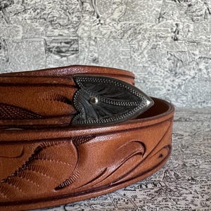May include: Close-up of a brown leather belt with a decorative silver buckle. The belt features embossed floral patterns and a detailed buckle design. The leather has a rich, warm tone, and the buckle is a contrasting silver color.