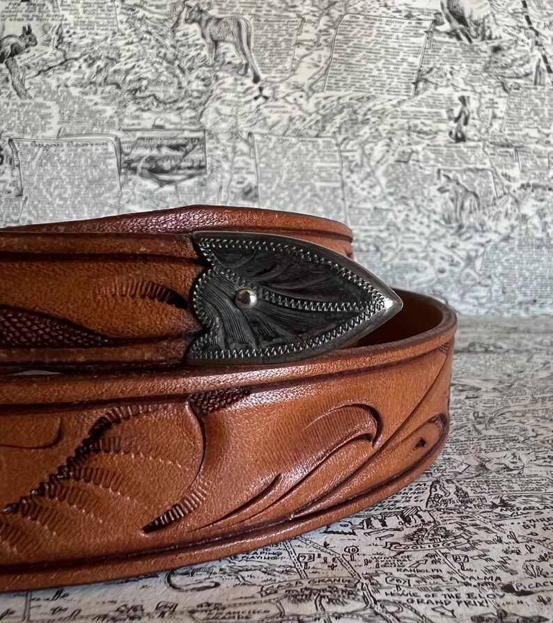 May include: Close-up of a brown leather belt with a decorative silver buckle. The belt features embossed floral patterns and a detailed buckle design. The leather has a rich, warm tone, and the buckle is a contrasting silver color.