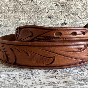 May include: A rolled brown leather belt with a decorative floral pattern. The belt has a metal buckle and several holes for adjustment. The leather is a warm, medium brown color, and the design is embossed into the surface.