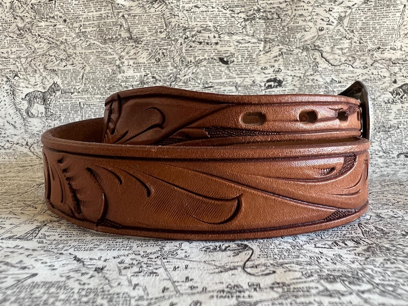 May include: A rolled brown leather belt with a decorative floral pattern. The belt has a metal buckle and several holes for adjustment. The leather is a warm, medium brown color, and the design is embossed into the surface.