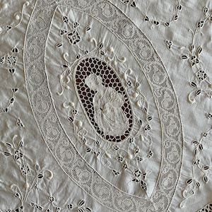 May include: White cotton tablecloth with intricate lace embroidery. The centre features a large oval with a delicate floral design and a smaller oval with a figure in the centre.